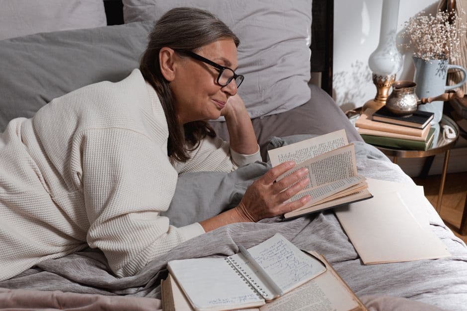 An older woman reading in bed under a blanket.